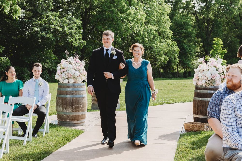 Couple walking down outdoor wedding aisle flanked by floral arrangements on barrels, guests seated on white chairs.