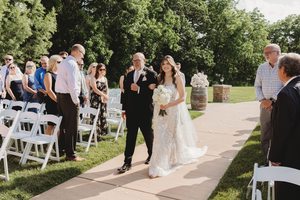 Bride walks down outdoor aisle with escort past white chairs at a sunny garden wedding ceremony.