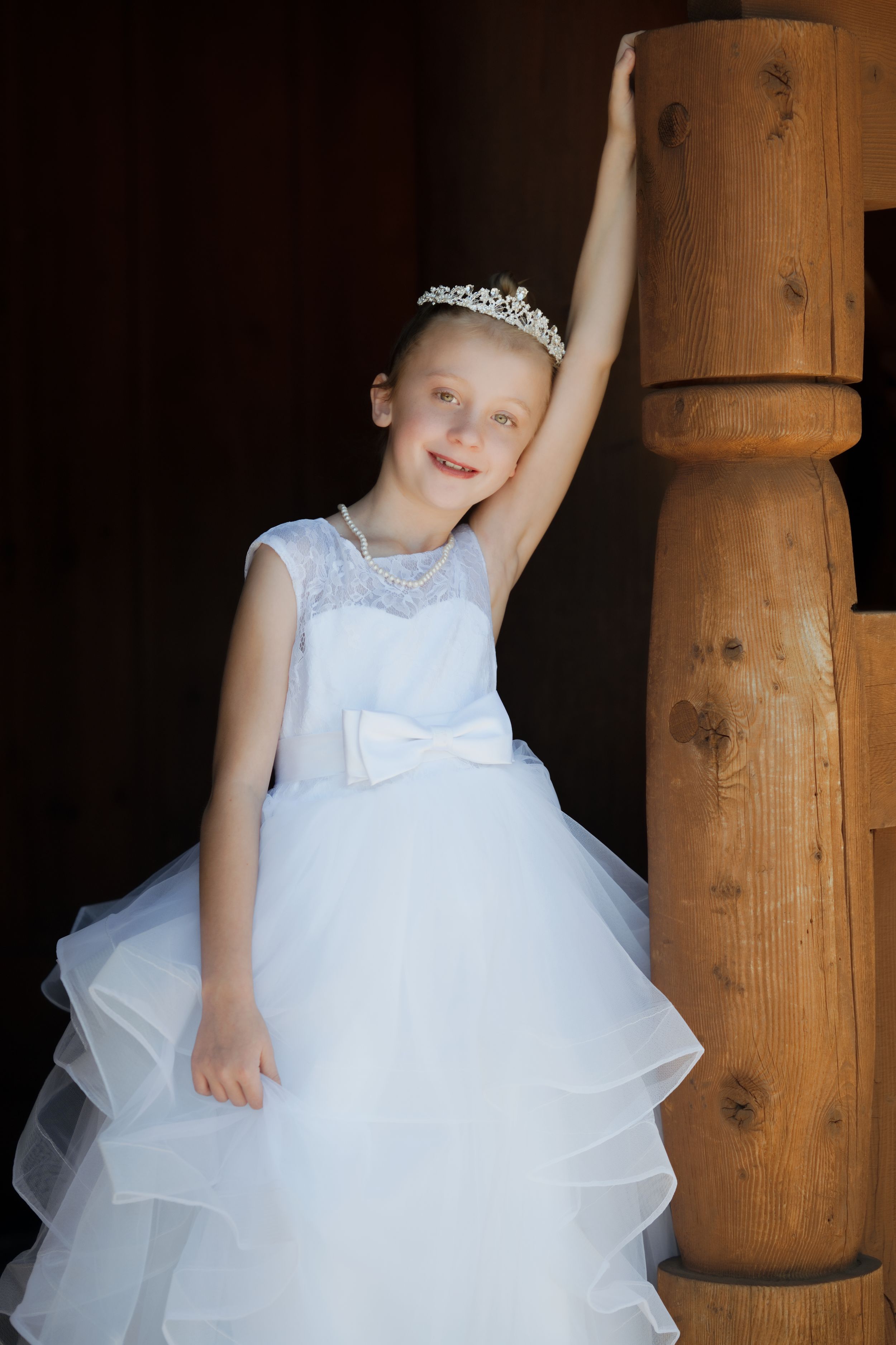 Young girl in white tiara and ruffled ball gown dress poses by wooden staircase post.