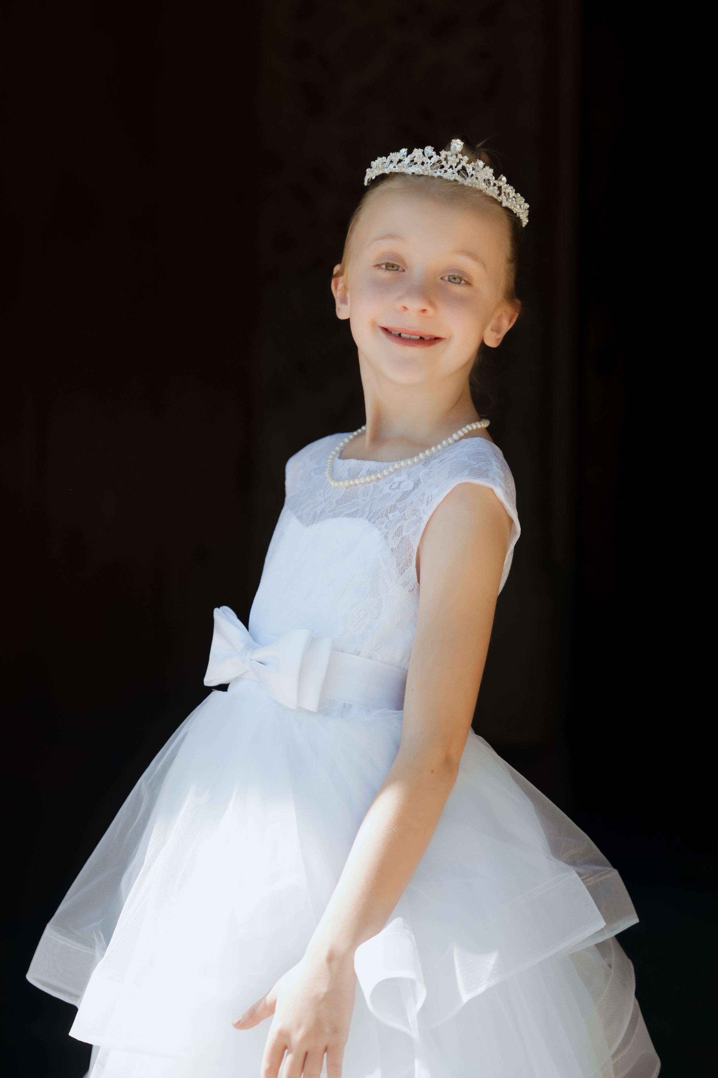 Young girl in white tiara and elegant white dress smiles joyfully against dark background.