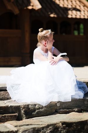 Young girl in white tulle dress and tiara sits on stone steps reading a book outdoors near wooden structure.