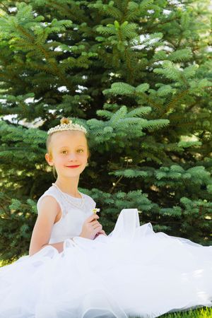 Young girl in white dress and tiara crown sitting outdoors in front of green evergreen trees.