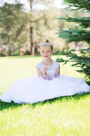 Young girl in white dress and tiara sits on green grass near evergreen tree, smiling for First Communion photo.
