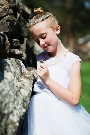 A young girl in a white dress and tiara holds a daisy while leaning against a stone wall outdoors.