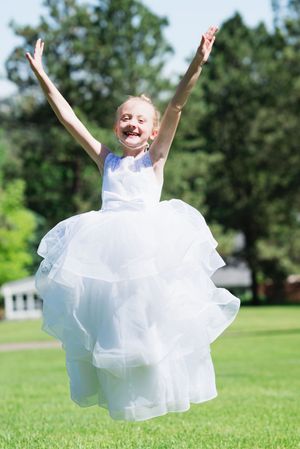 A joyful young girl in a white tulle dress jumps with arms raised on a sunny green lawn.