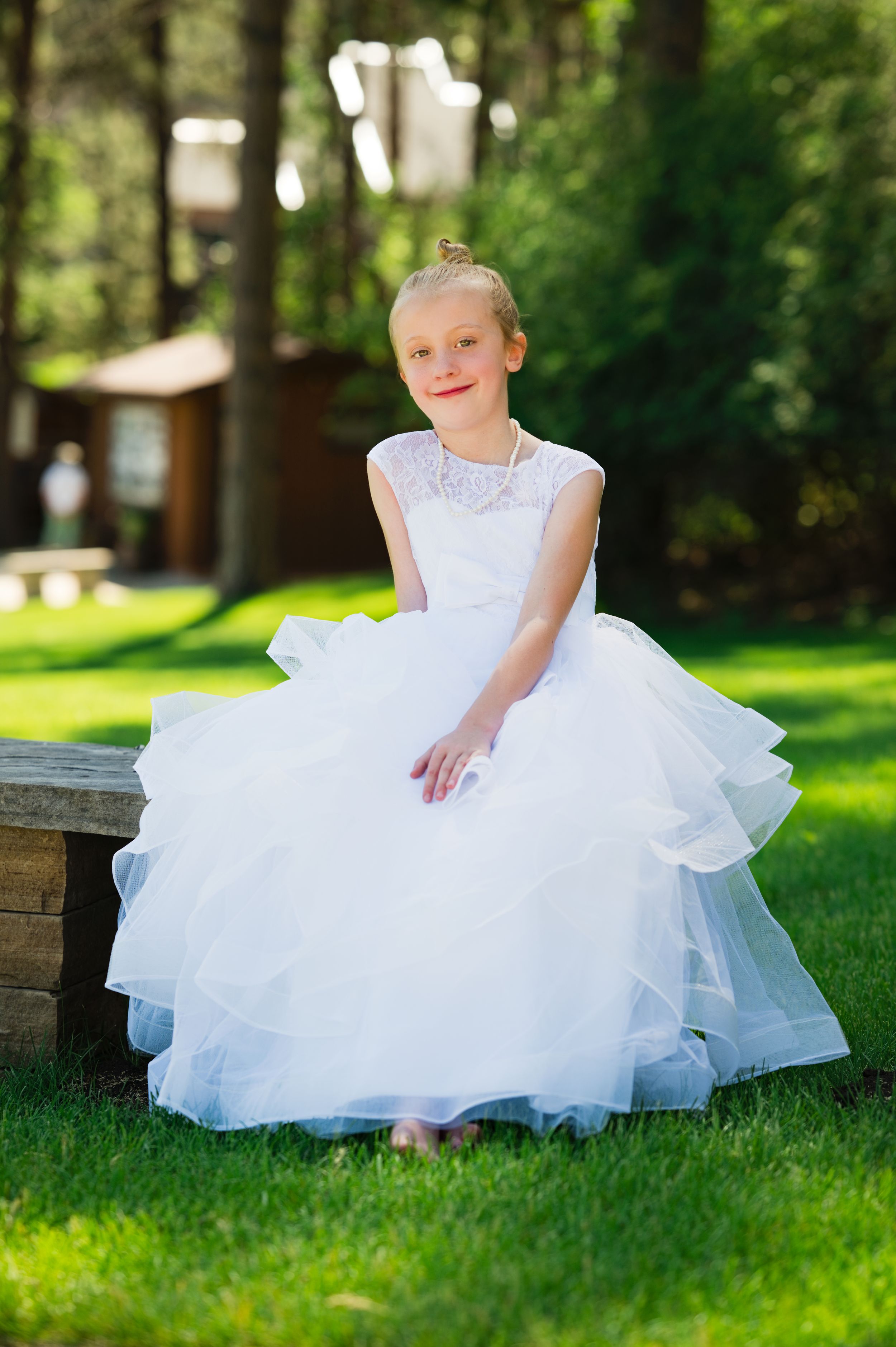 Young girl in white lace ball gown dress sitting outdoors on stone bench surrounded by green grass and trees.