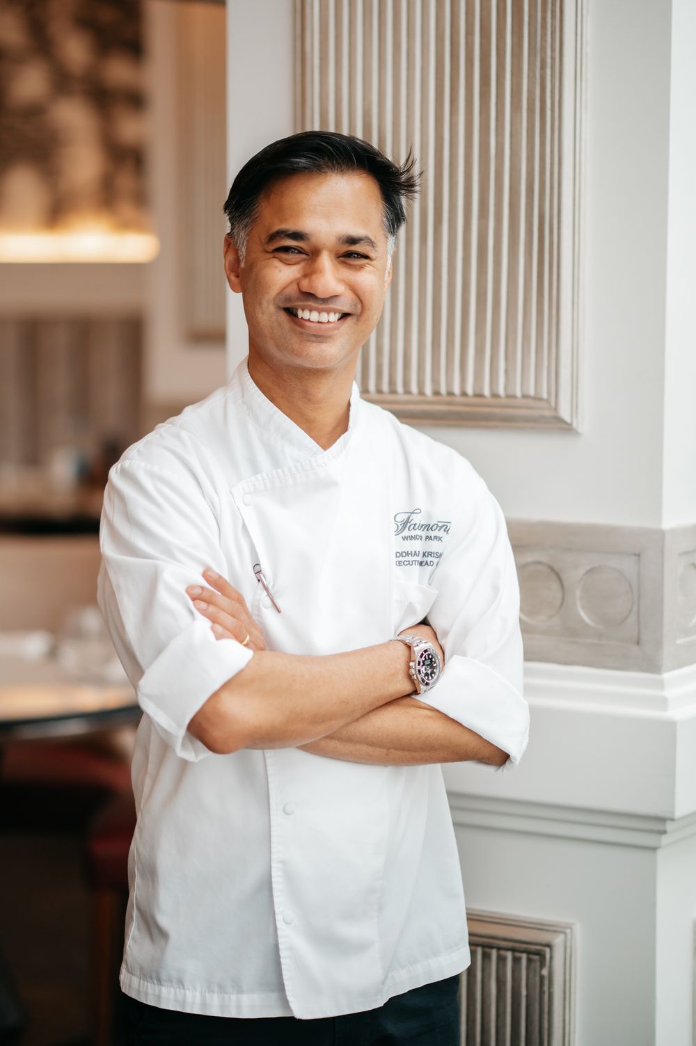 Smiling chef in white uniform with arms crossed standing in an elegant restaurant dining room.