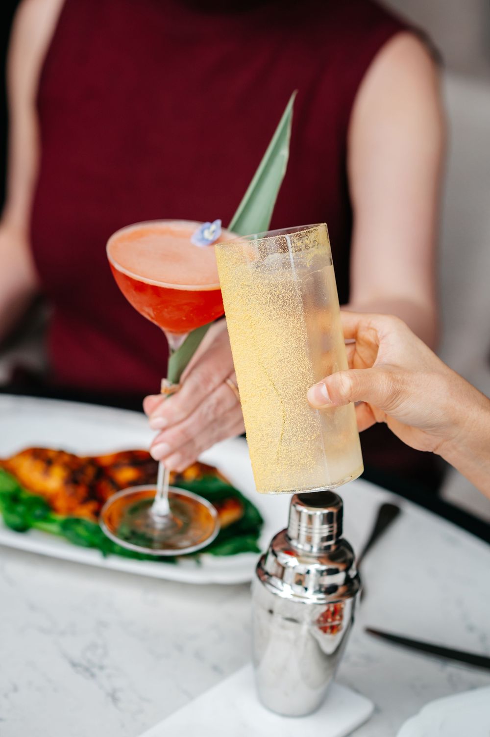 Two hands clinking cocktails at a restaurant table, one pink martini and one sparkling highball with pineapple garnish.