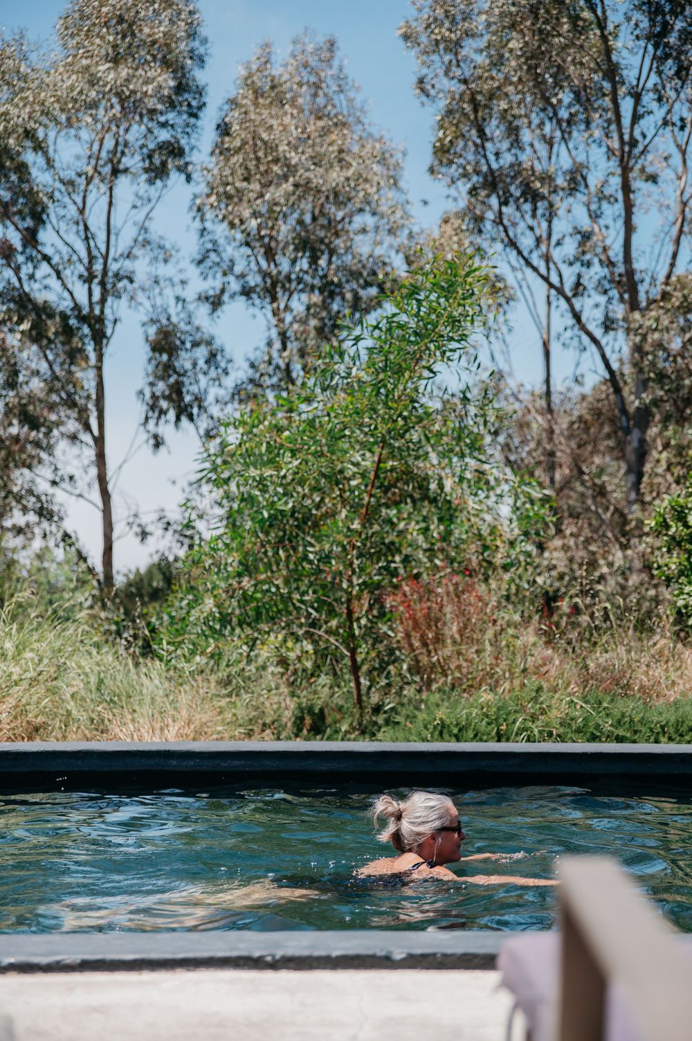 Person relaxing in an outdoor pool surrounded by lush green trees and tall grass on a sunny day.