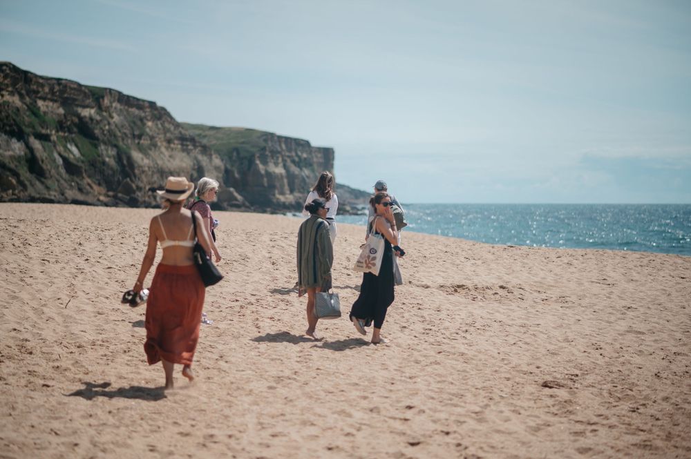 Four women walking on a sandy beach with dramatic cliffs and blue ocean in the background on a sunny day.