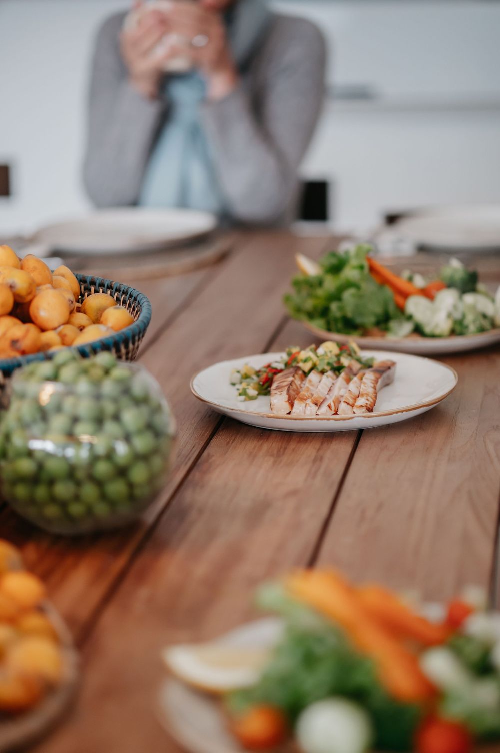 Wooden dining table with fresh vegetables, fruits, and appetizer plates, with a blurred person sitting in the background.