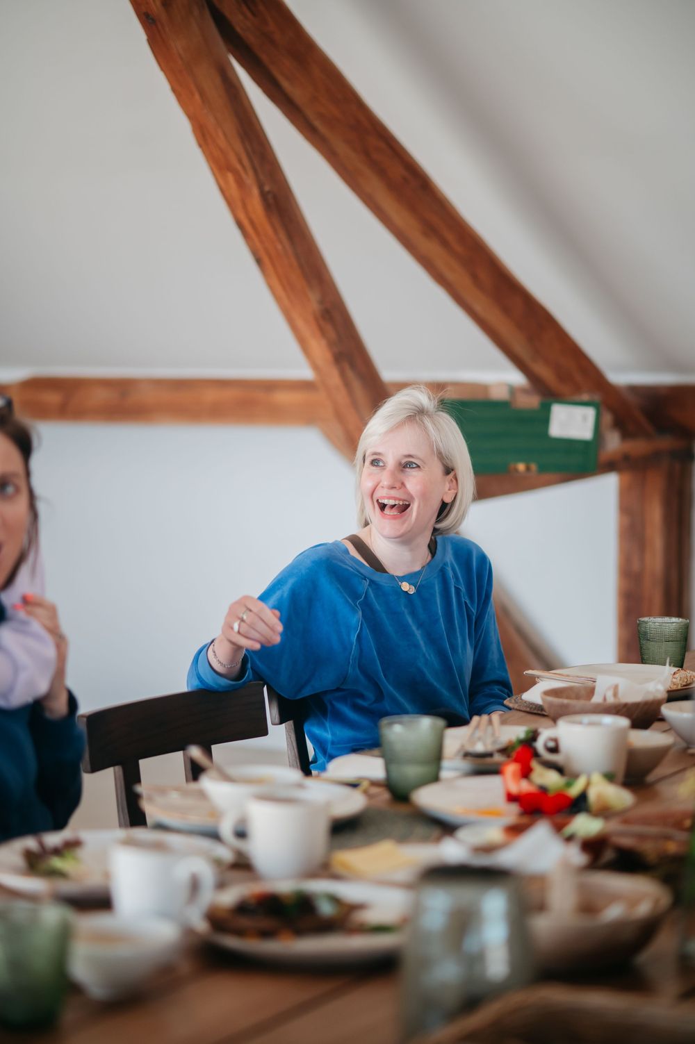 A smiling woman in a blue top laughs joyfully at a rustic dining table set with food and drinks.