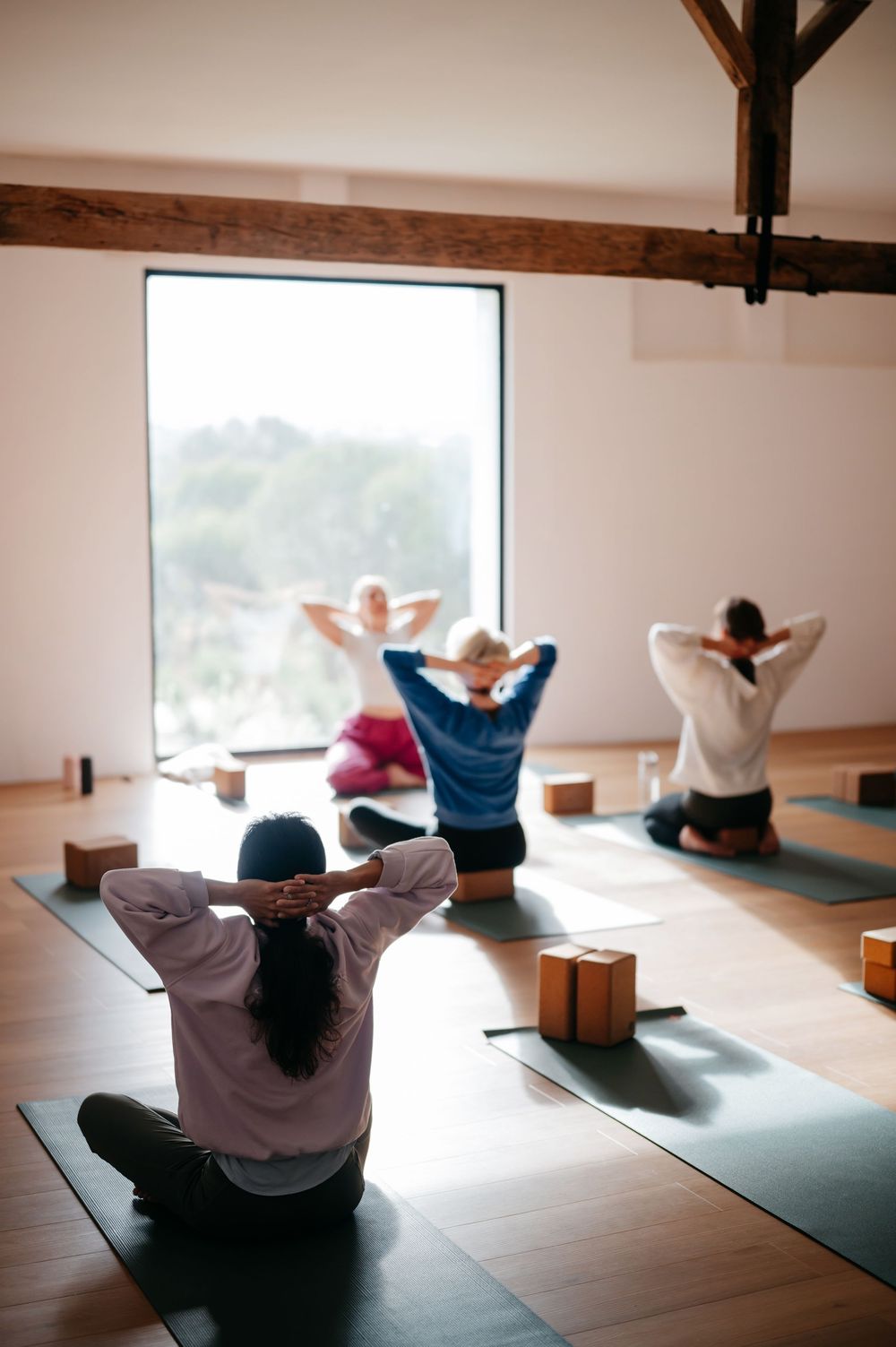 Indoor yoga class with students sitting cross-legged, hands behind heads, on mats with wooden blocks in bright studio.