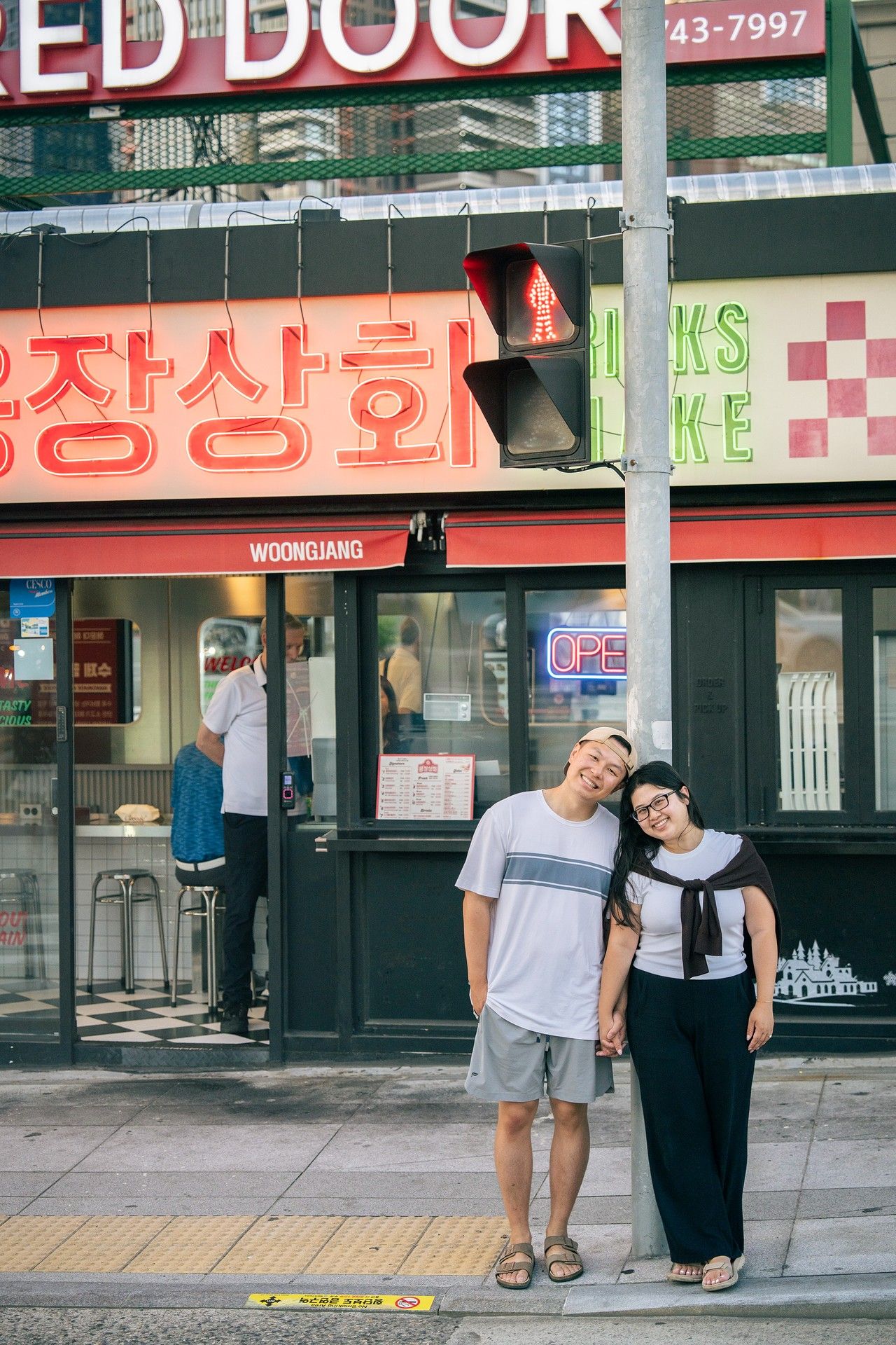 A couple smiles and poses together on a sidewalk outside Woong Jang Korean store with neon signs in Koreatown.