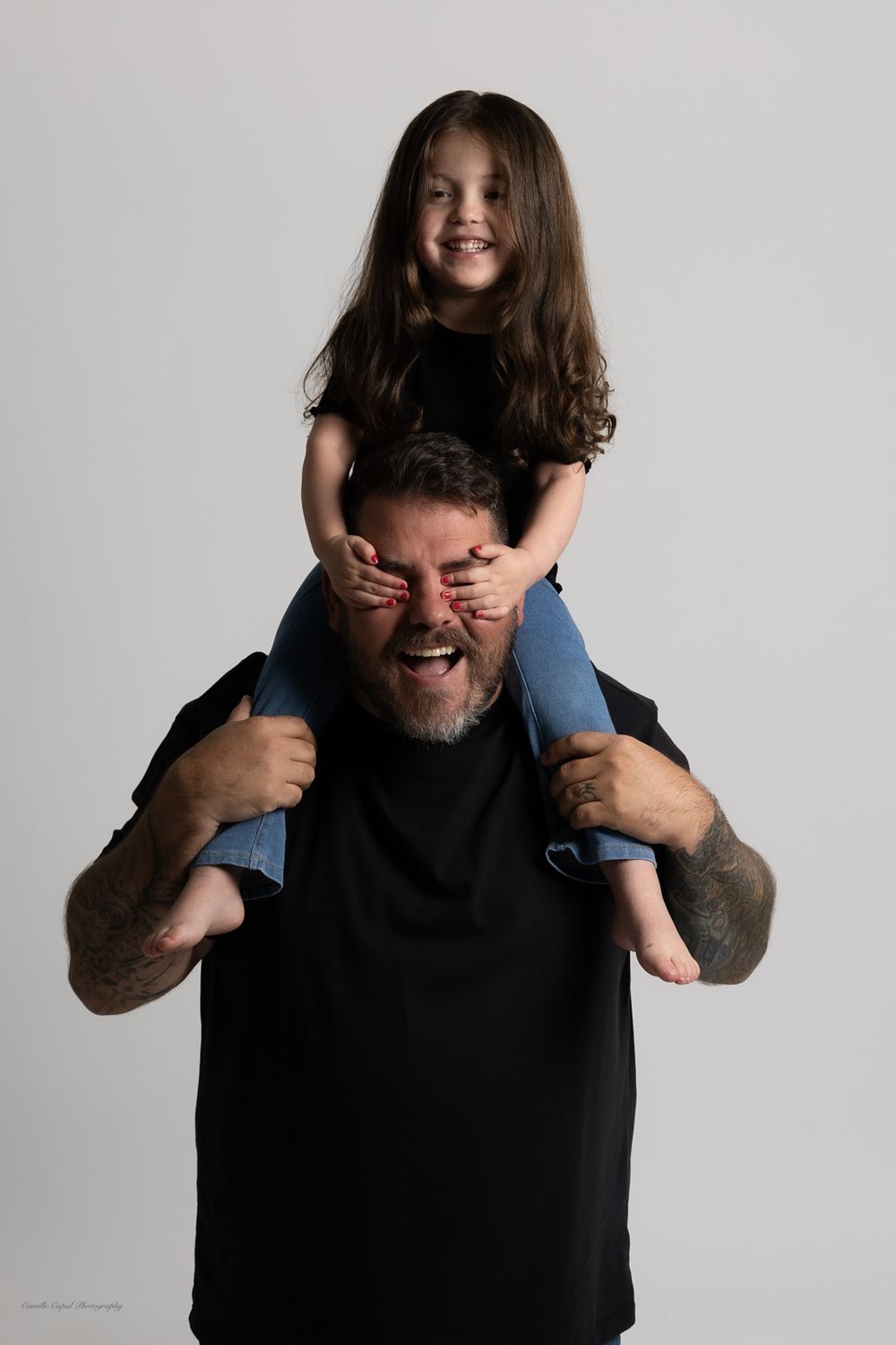 A young girl sits on a dads shoulders, covering his eyes and smiling in a studio portraitfamily portrait session in Pittsburgh by Cupid