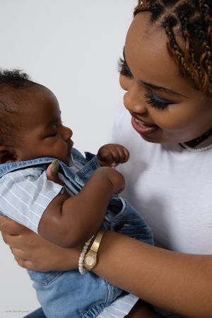 A young Black mother smiles lovingly at her newborn baby dressed in a denim outfit against a white background family portrait session in Pit