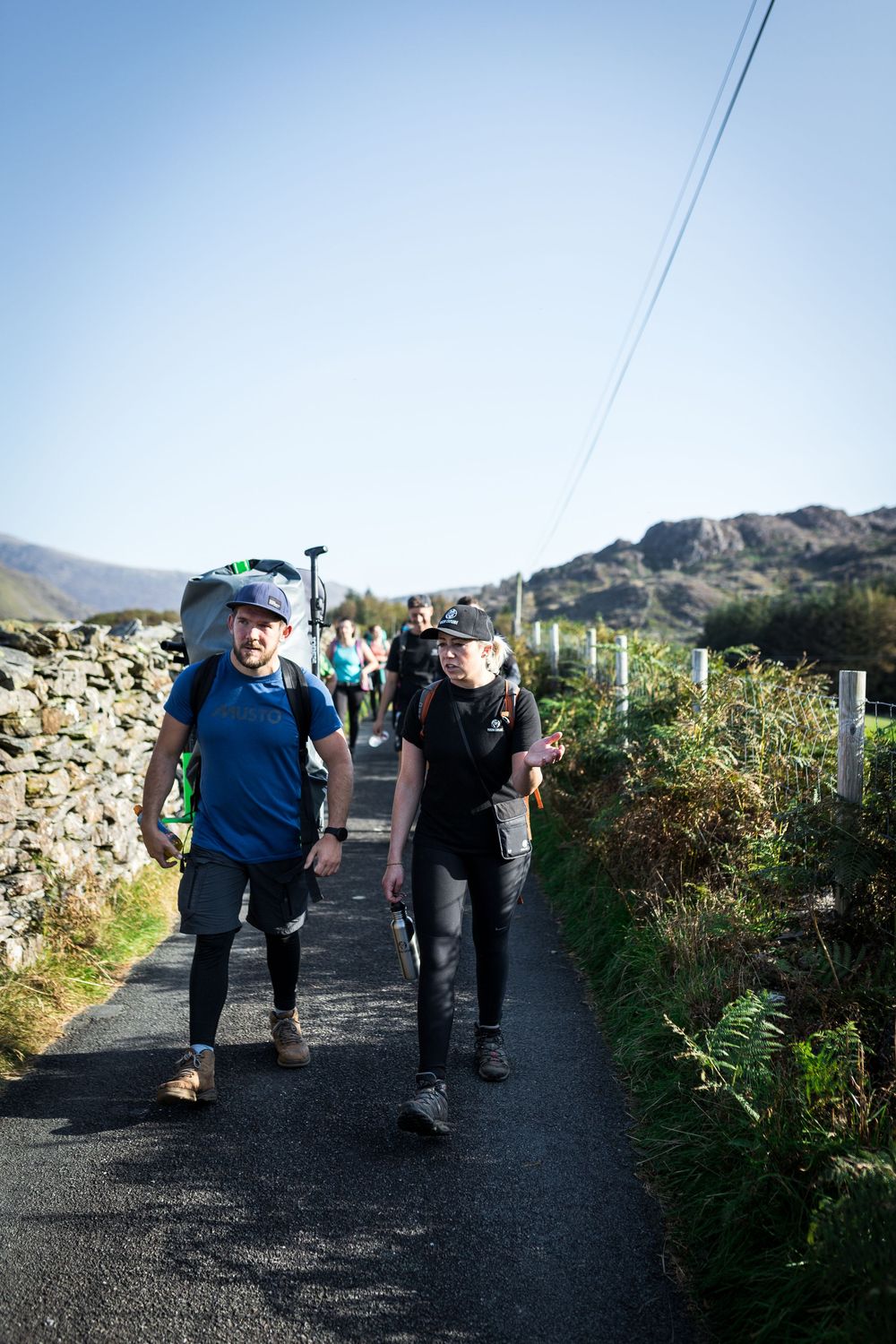 Hikers walking along a narrow rural path with stone walls and mountains in the background on a clear day.
