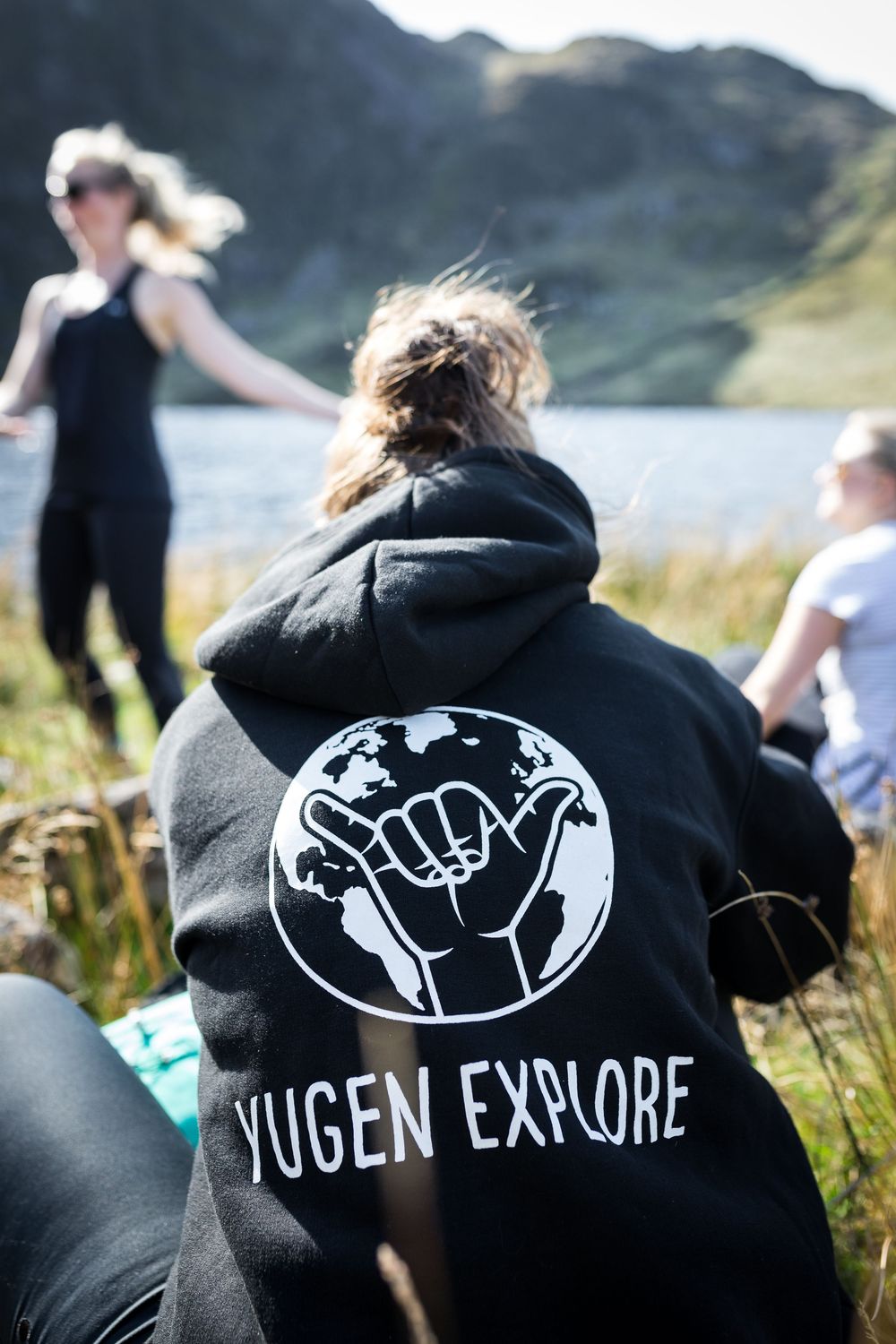 Person wearing a black Yugen Explore hoodie with globe logo, sitting outdoors near a mountain lake.