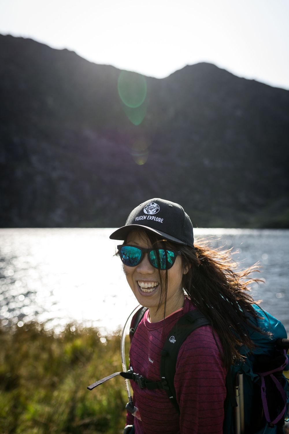 Smiling woman hiker with backpack, cap, and mirrored sunglasses standing near a mountain lake in bright sunlight.