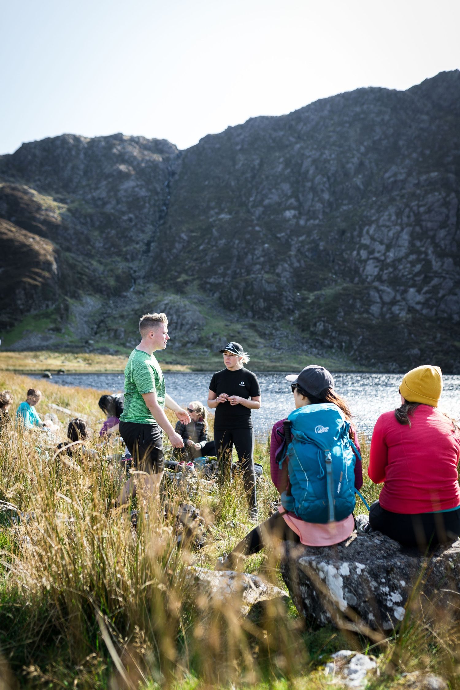 Group of hikers resting by a mountain lake on a sunny day, surrounded by rocky peaks and golden grass.