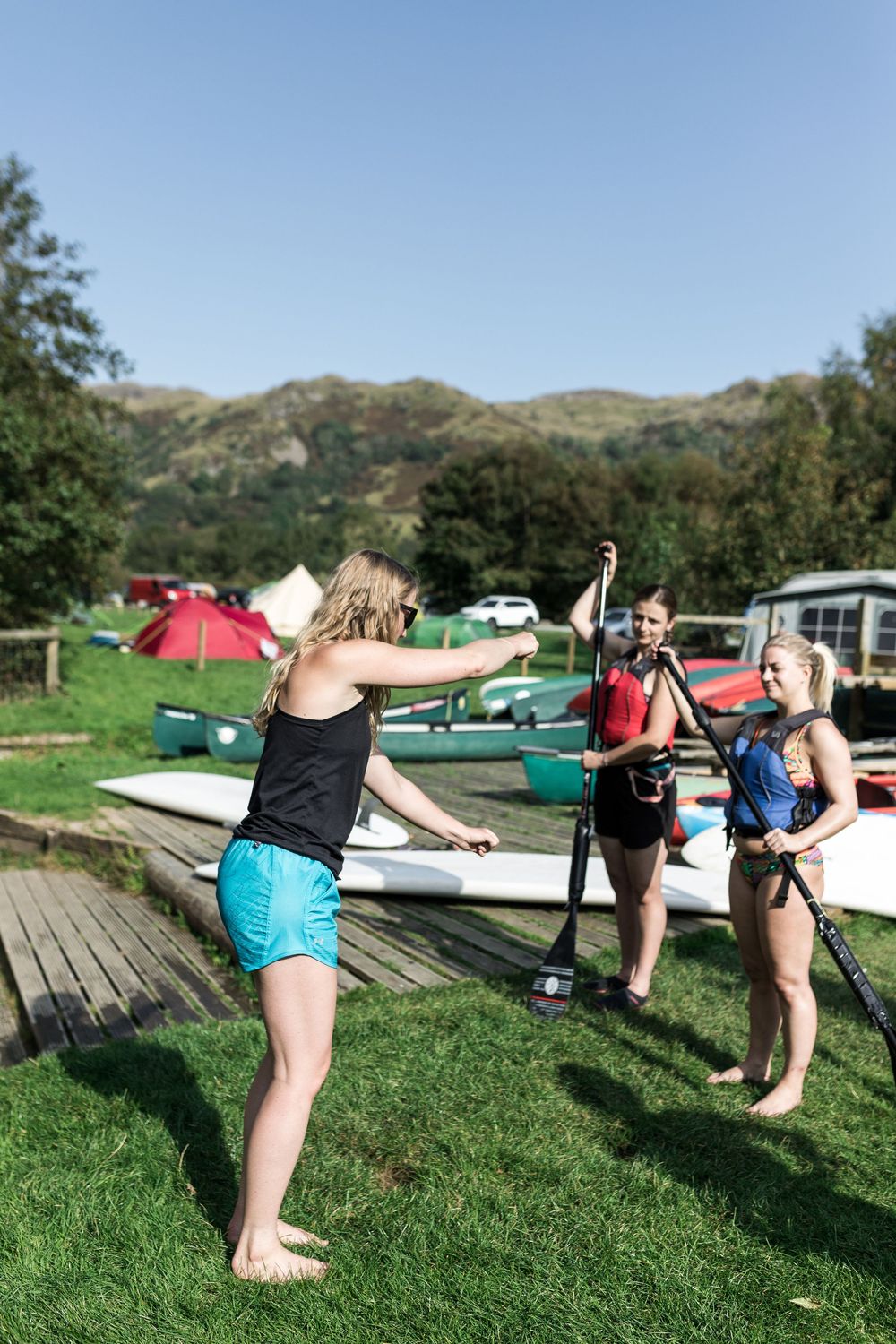 Three women practicing paddleboard techniques on grass near a lakeside outdoor activity center.