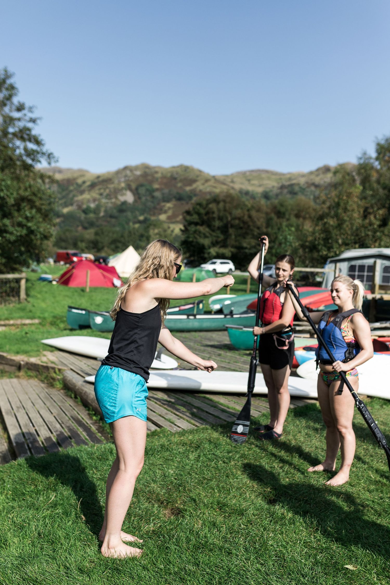 Three women practicing paddleboard techniques on grass near a lakeside outdoor activity center.