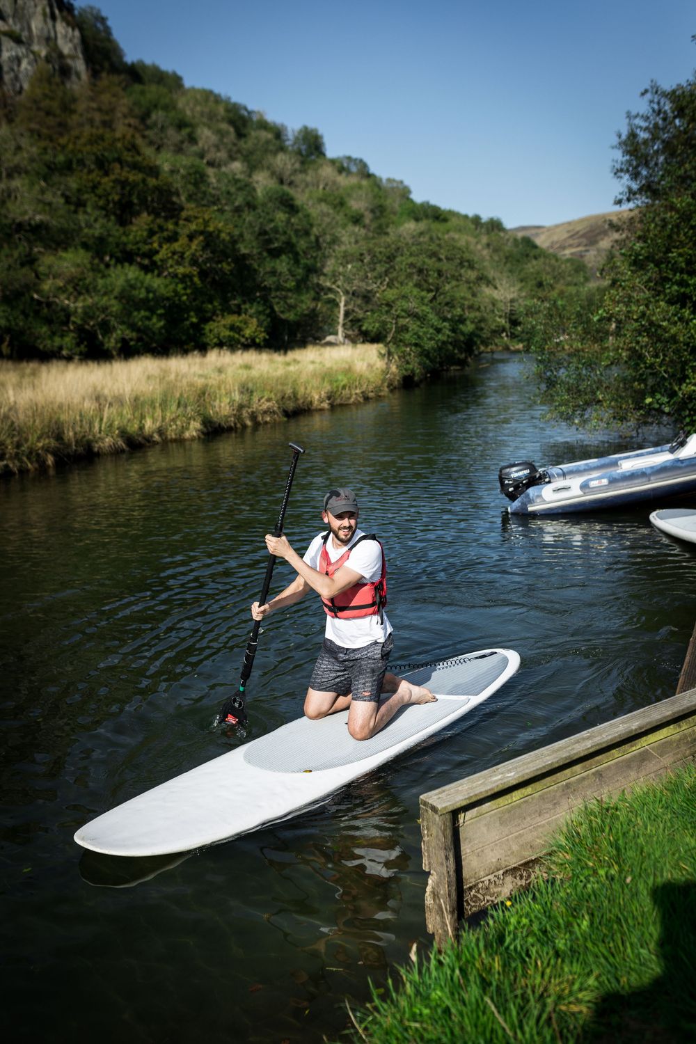 Man jumping on a white stand-up paddleboard on a calm river surrounded by lush green trees.