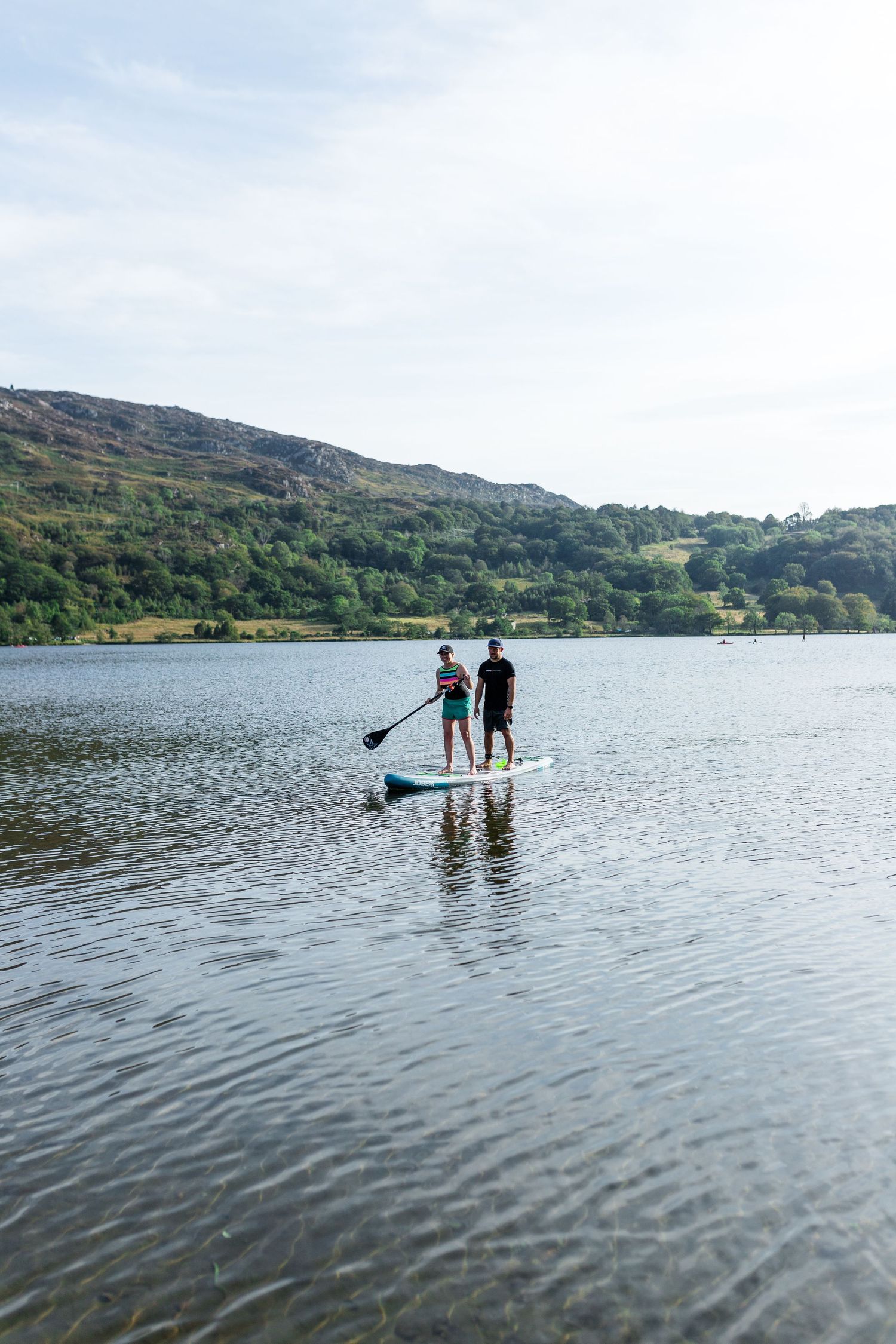 Two people stand-up paddleboarding together on a calm lake with green hills in the background.