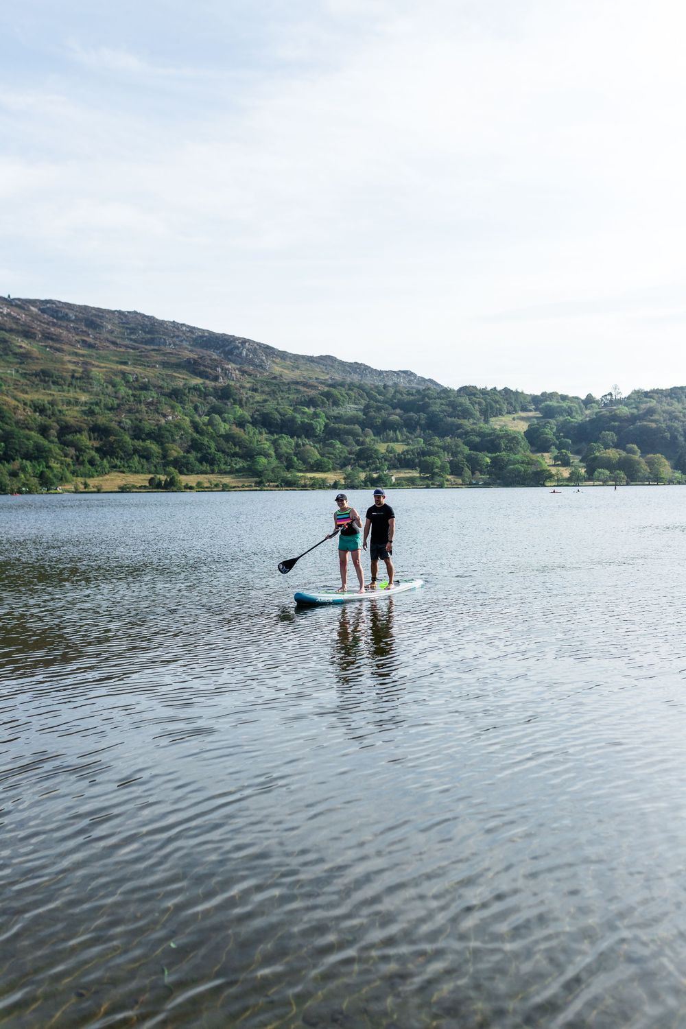 Two people stand-up paddleboarding together on a calm lake with green hills in the background.