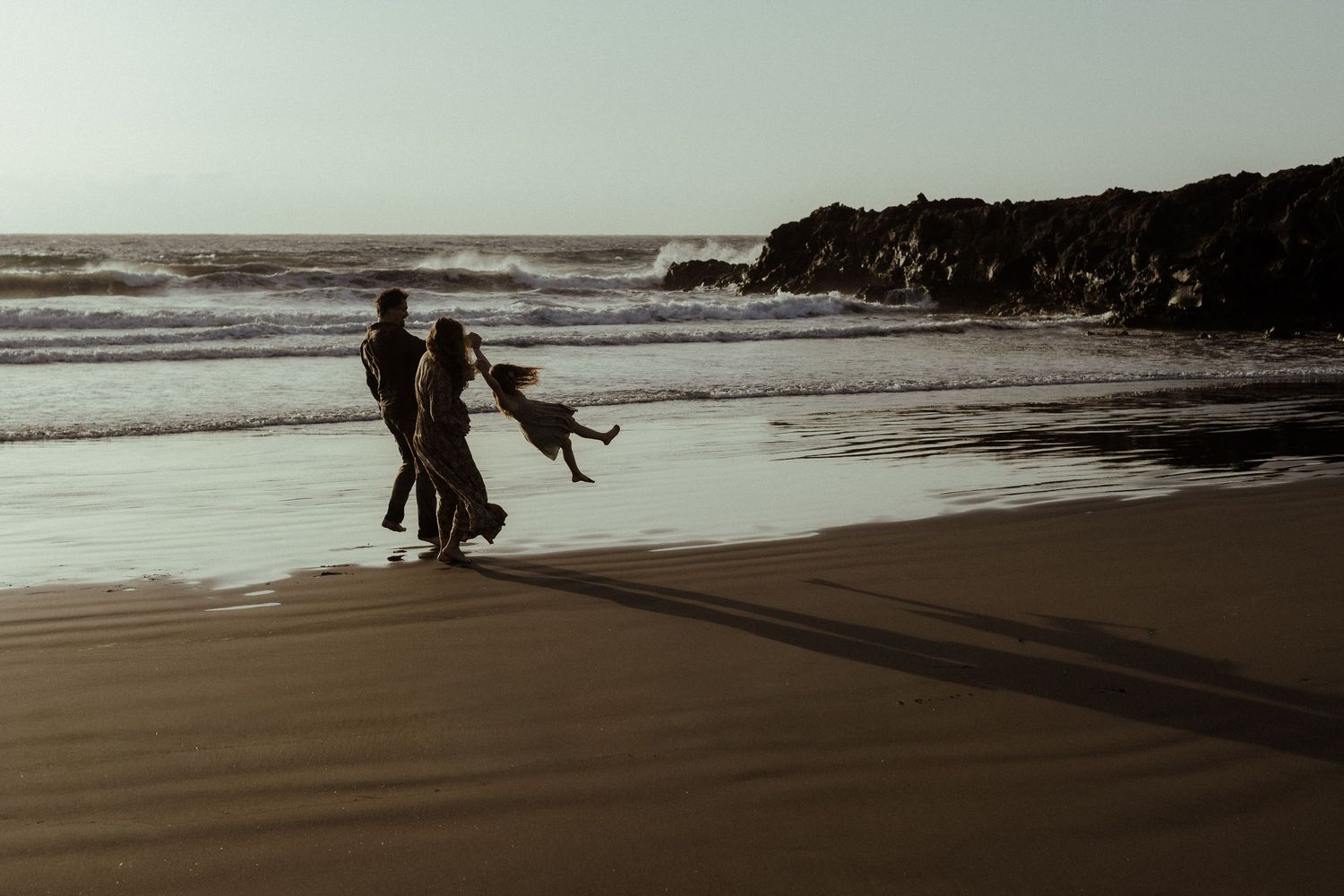 Couple dancing on a sandy beach at sunset with ocean waves and rocky cliffs in the background.