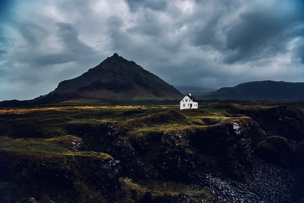 A lone white house sits beneath a dramatic volcanic mountain peak under stormy skies in Iceland.