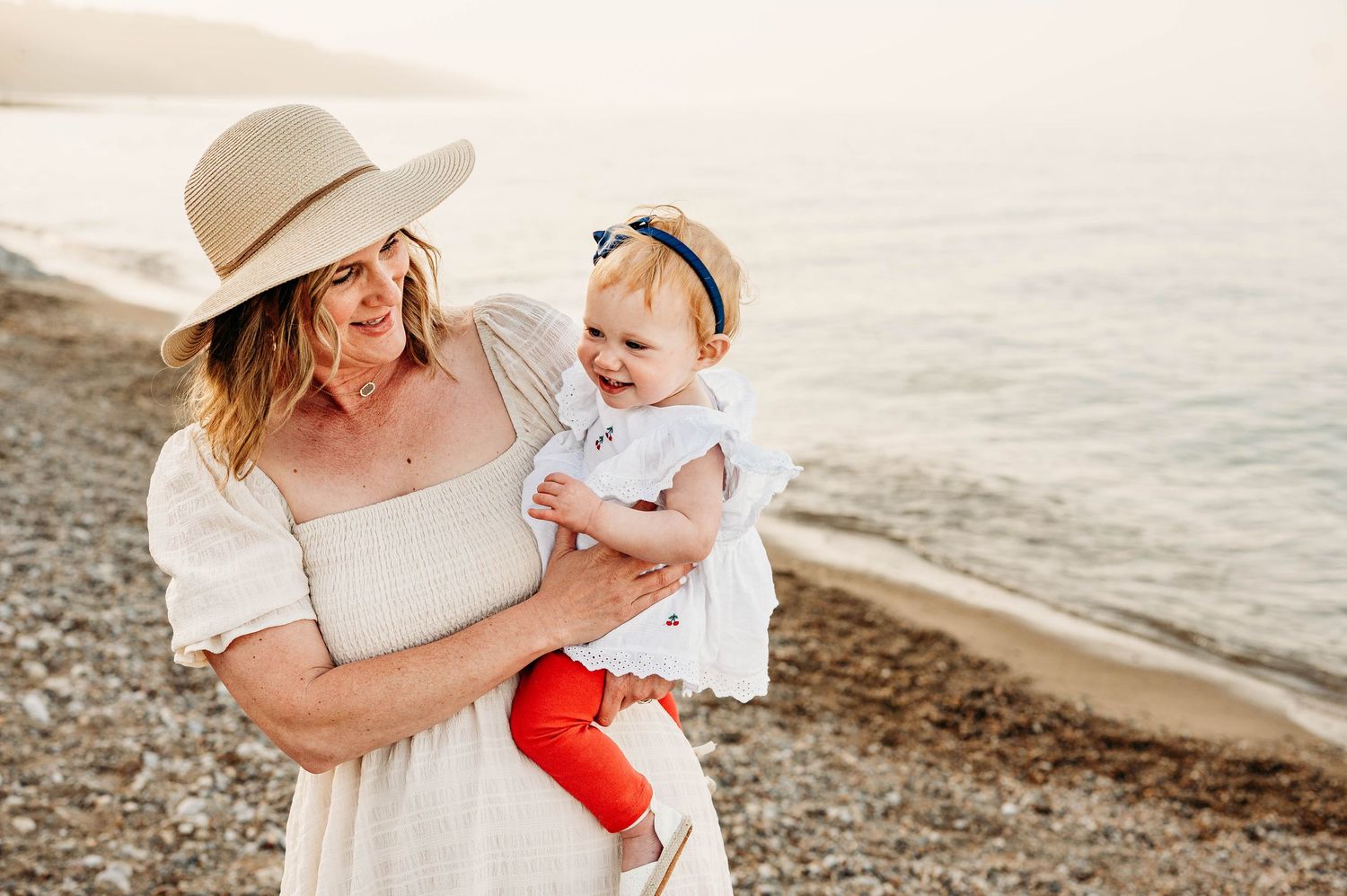 Woman in white dress and hat holds smiling baby girl on a rocky beach at sunset.