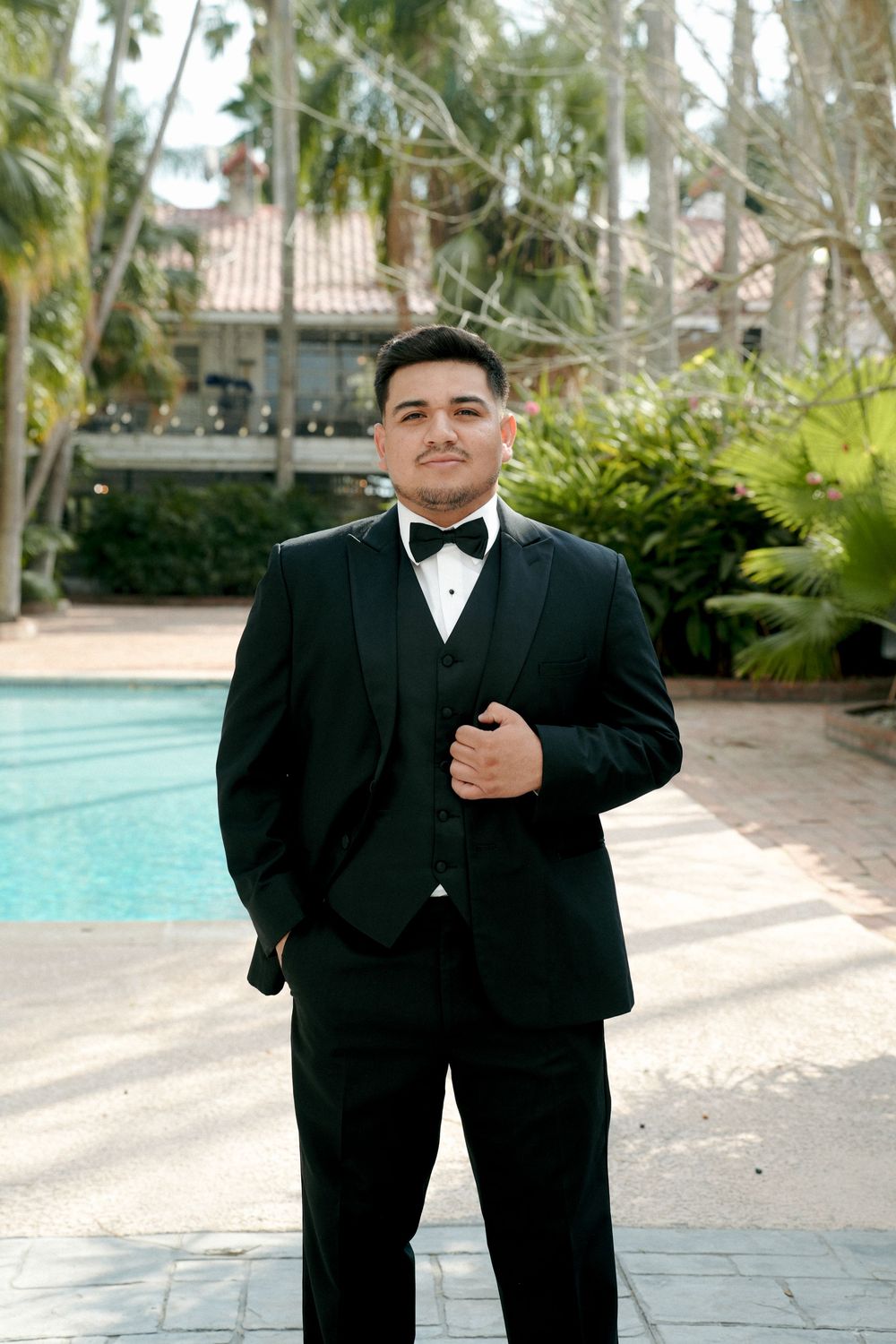 Young man in black tuxedo with bow tie posing poolside at an elegant outdoor venue surrounded by palm trees.