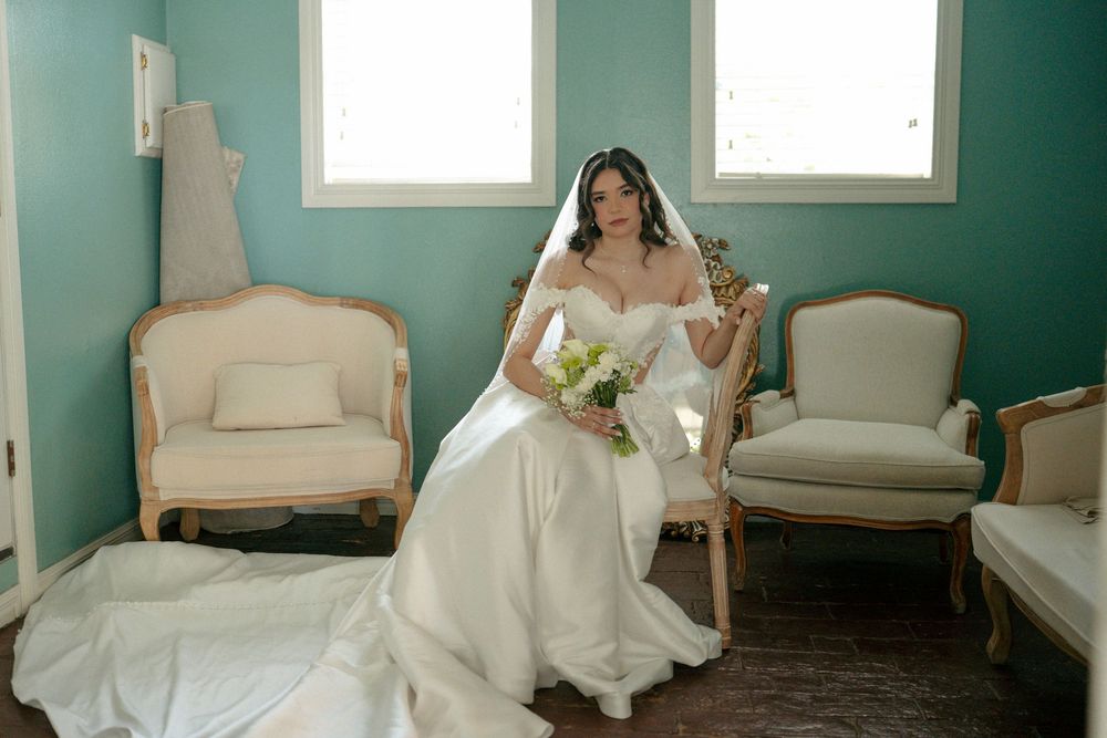 Bride in white off-shoulder gown with veil holds bouquet, seated in elegant teal room with French-style chairs.