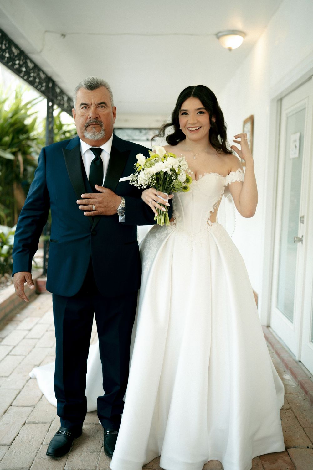 Bride in white ballgown holding bouquet walks with older man in dark suit down outdoor corridor.