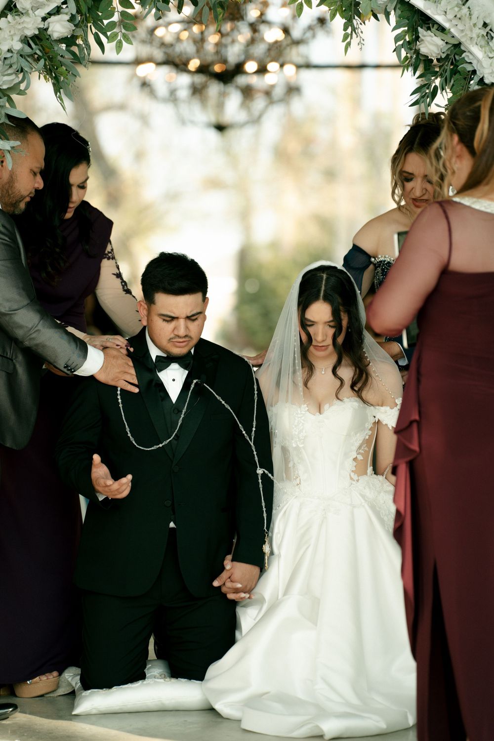 Bride and groom kneeling in prayer during wedding ceremony, surrounded by family members placing hands on them.