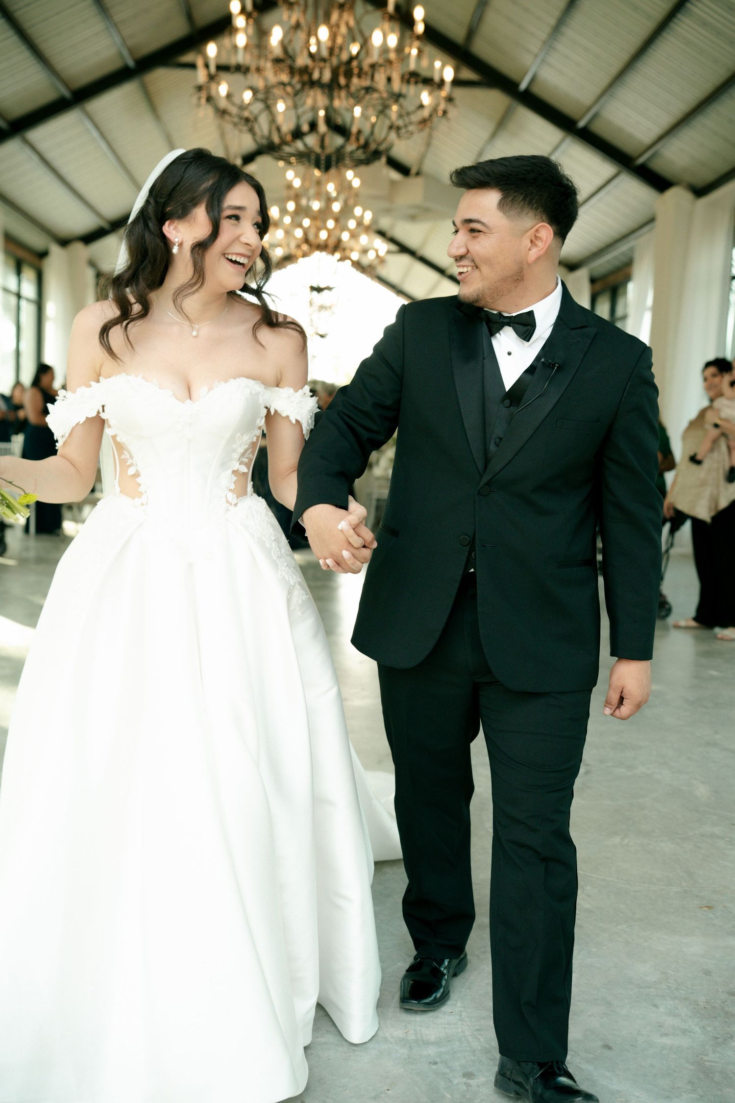 Bride in white off-shoulder gown and groom in black tuxedo hold hands and smile at each other at elegant wedding reception.