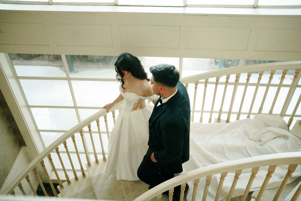 Bride and groom share a romantic moment on a white spiral staircase, bride in flowing white gown, groom in black suit.