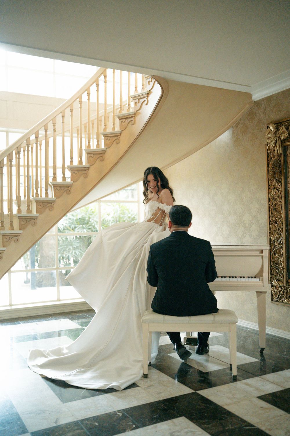 Bride in white gown stands by grand staircase as groom plays white piano in elegant marble-floored venue.