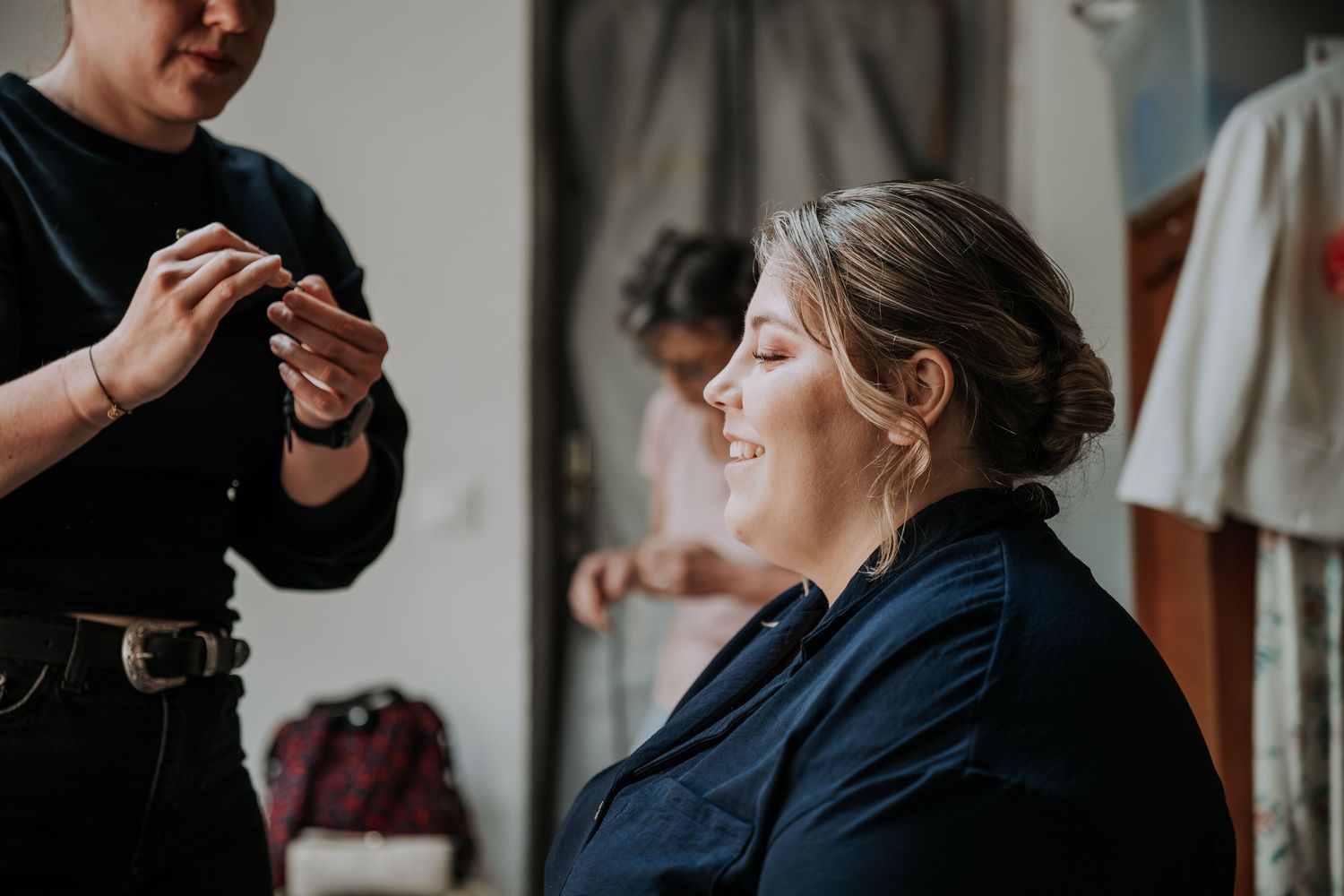 Bride smiling while getting hair styled by makeup artist before wedding ceremony indoors.