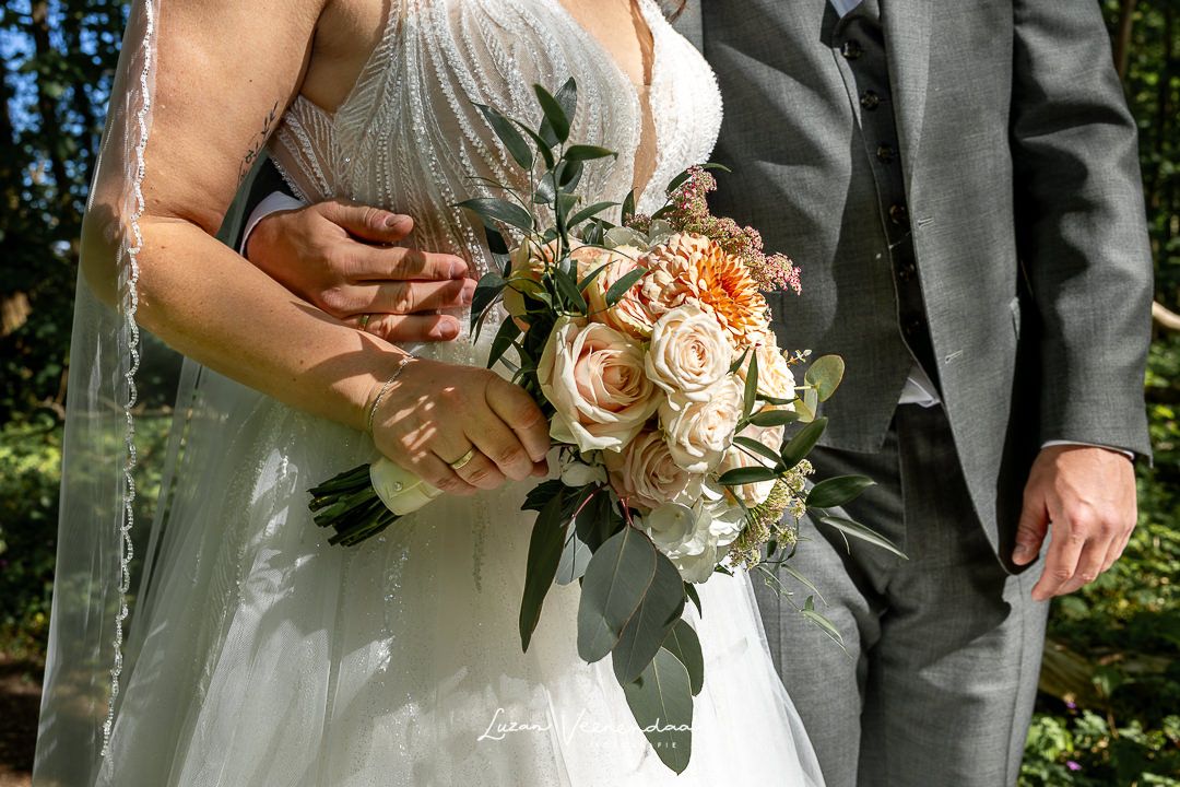 Detailfoto van het trouwboeket van Bloemore, trouwring aan de vinger en de arm van de bruidegom om de bruid geslagen bruiloft J&S