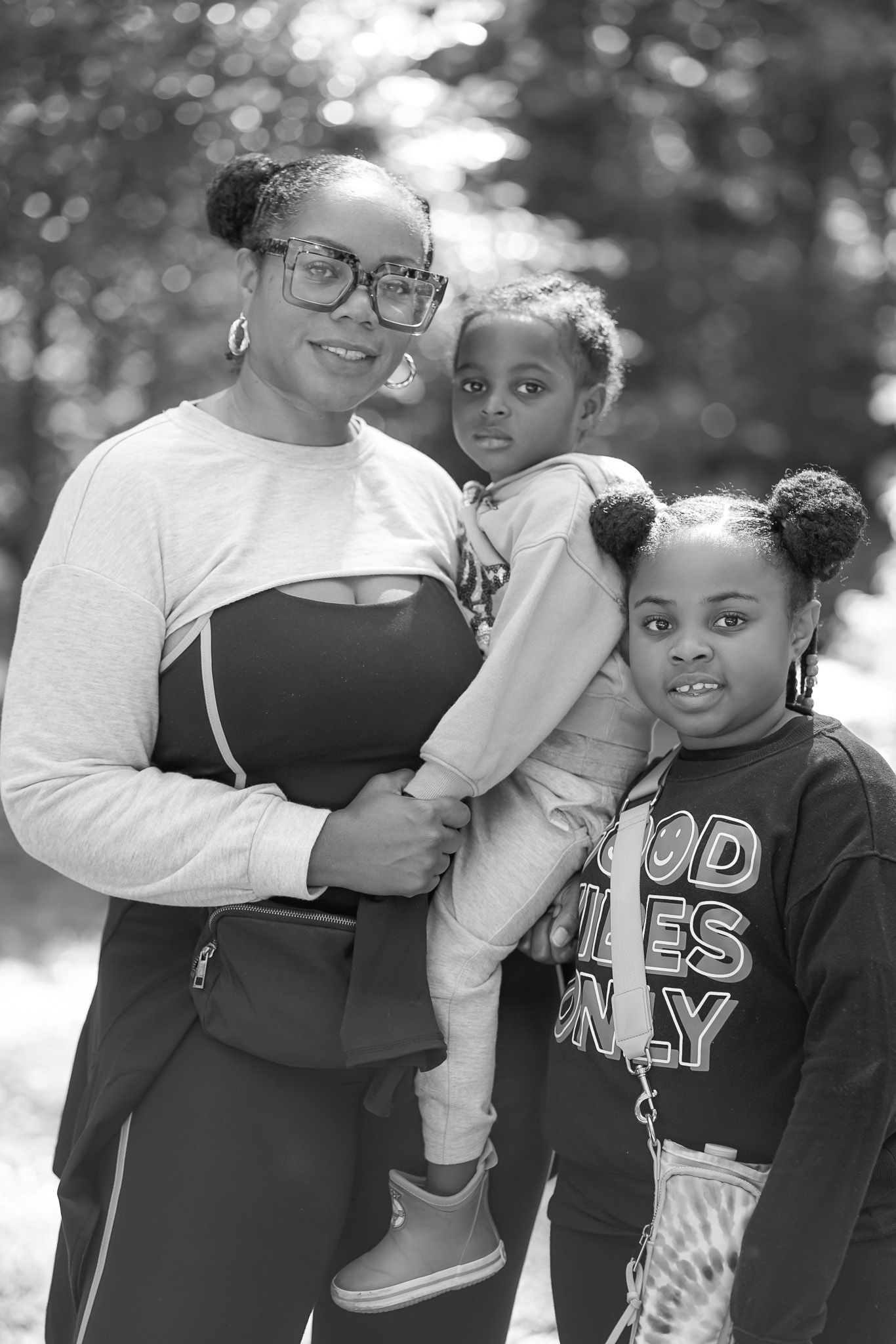 Black and white portrait of a mother holding two young daughters outdoors, smiling together near trees.