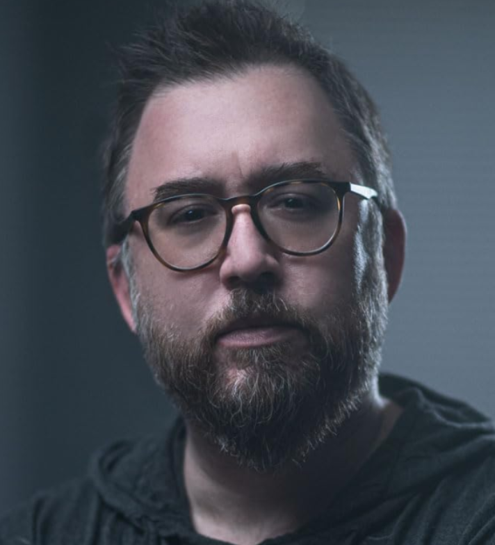 Close-up portrait of a bearded man wearing tortoiseshell glasses with a thoughtful expression.