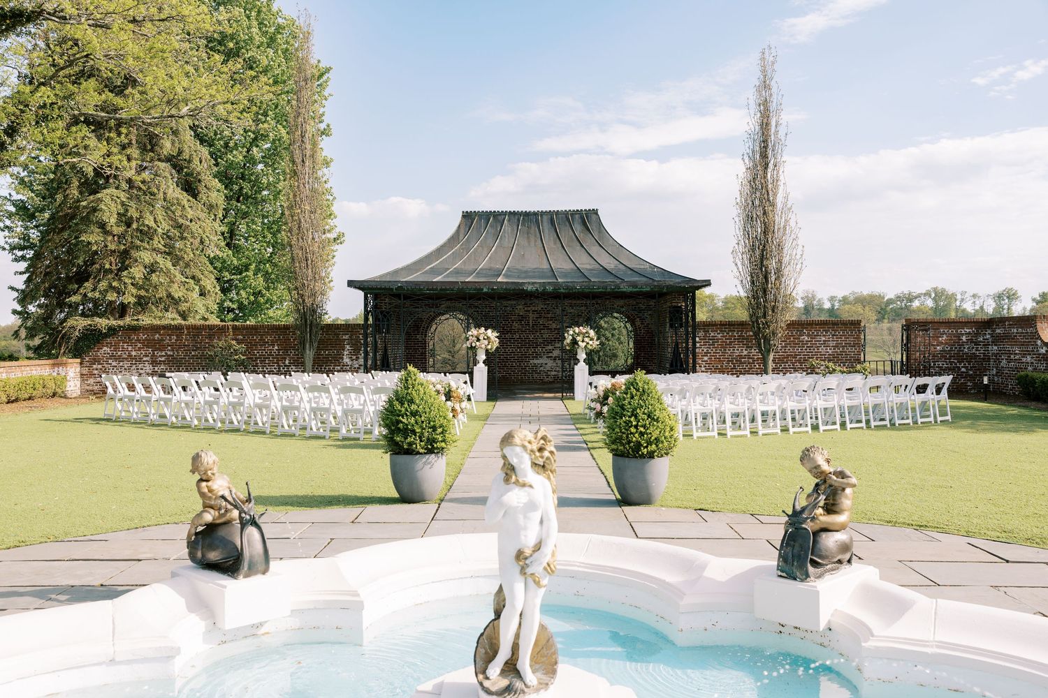Elegant outdoor wedding venue with white chairs, a pagoda gazebo, manicured lawn, and decorative fountain statues in a walled garden.