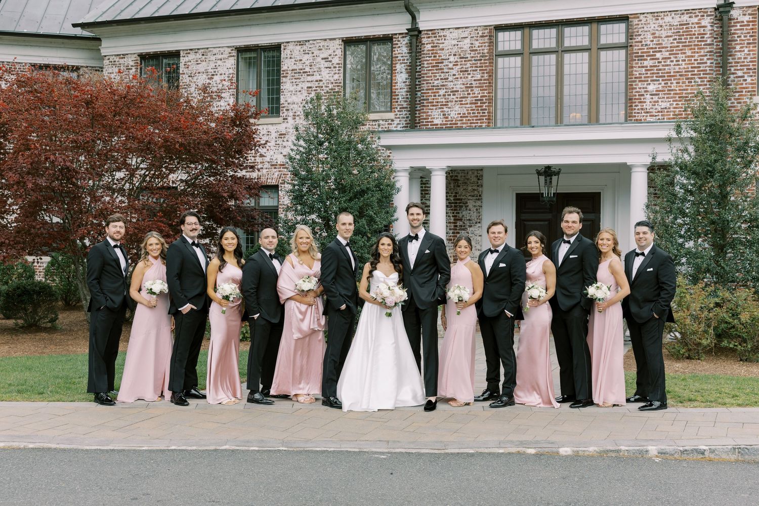 Wedding party posing outside elegant brick mansion, bridesmaids in pink gowns and groomsmen in black tuxedos flanking bride and groom.