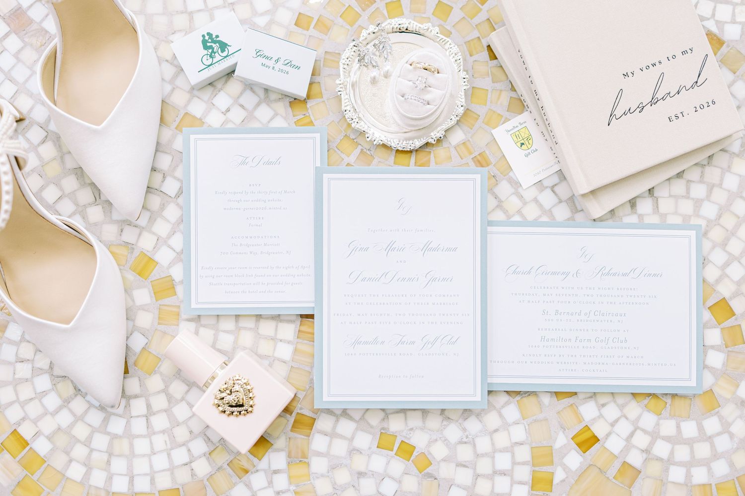 Elegant wedding stationery suite with light blue borders displayed alongside white heels and accessories on mosaic tile.