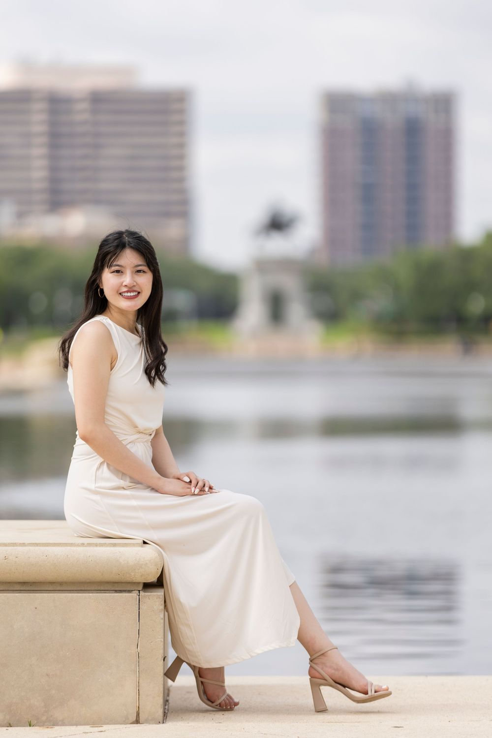 Young Asian woman in cream dress sitting by urban lake, smiling confidently with city skyline background.