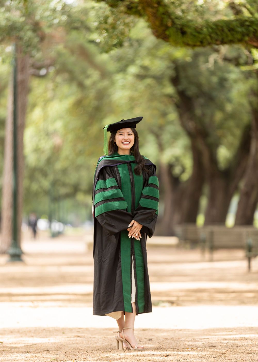 Woman in black and green doctoral graduation gown and cap smiling outdoors on a tree-lined path.