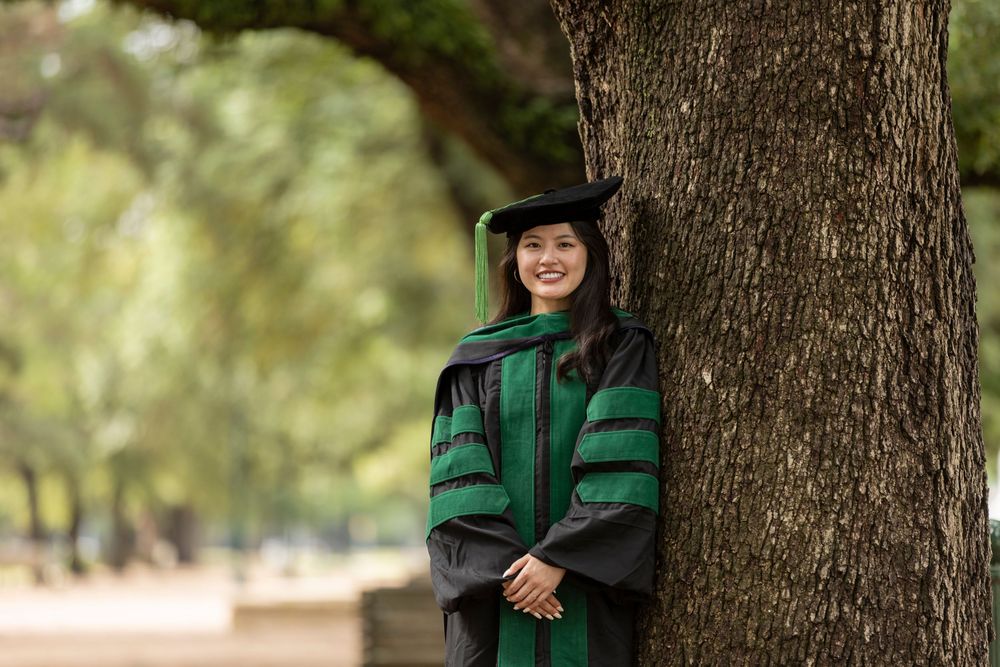 Woman in green doctoral graduation gown and cap smiling while leaning against a tree outdoors.