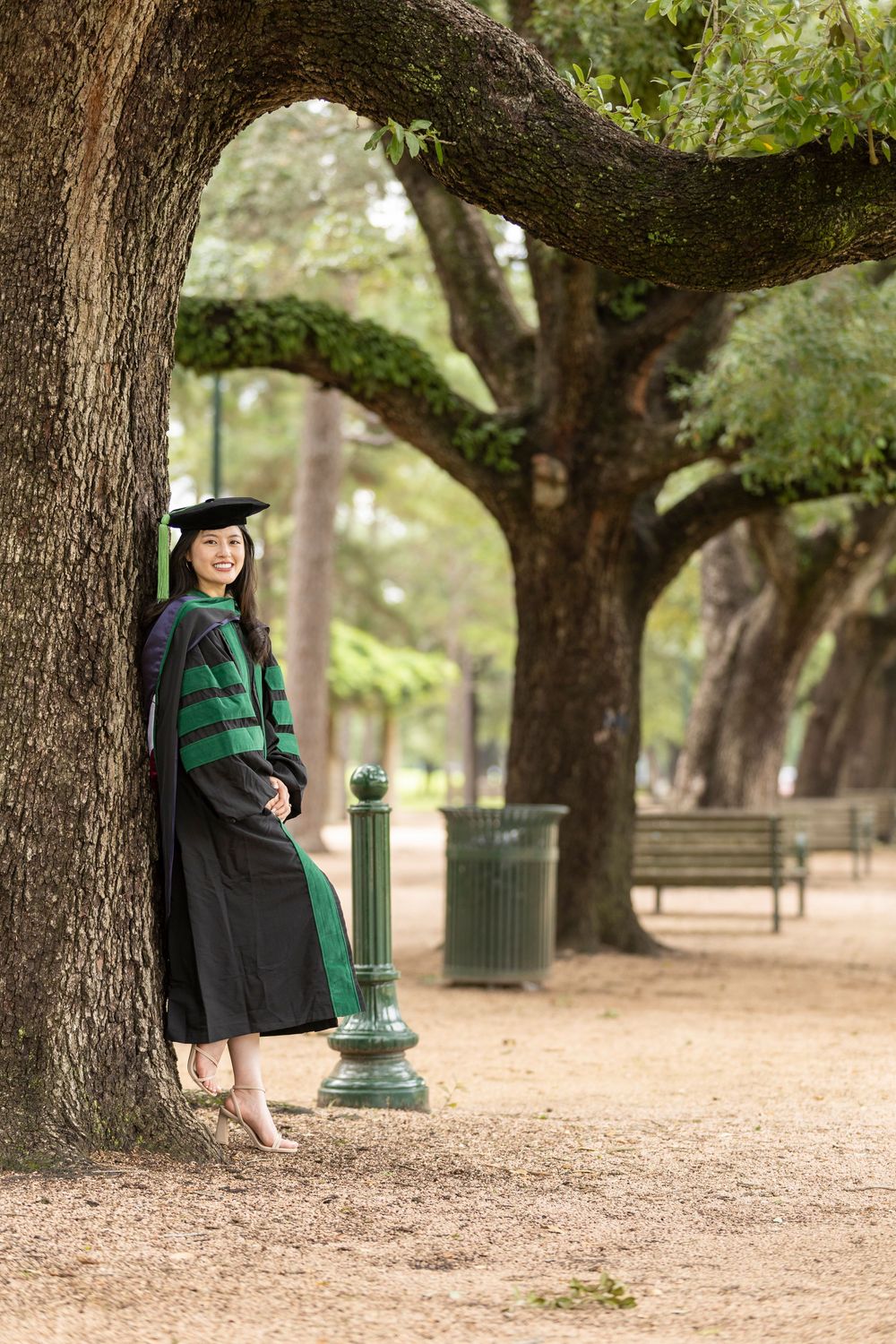 Woman in black and green doctoral graduation regalia poses under large oak trees in a park setting.